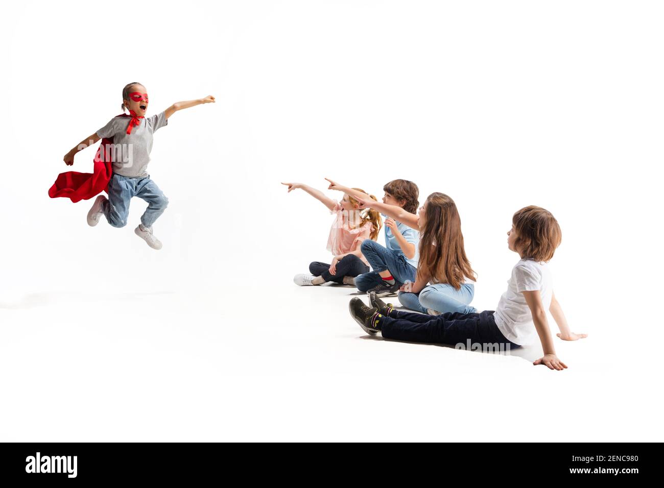 Flying. Child pretending to be a superhero with his friends sitting around  him. Kids excited and inspired by their brave friend in red coat isolated  on white background. Dreams, emotions concept Stock