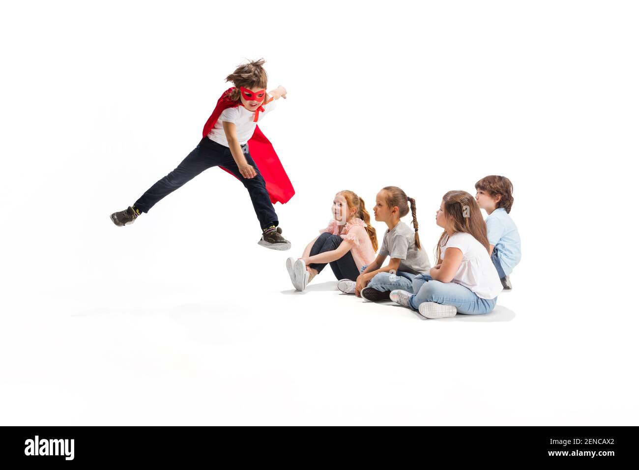 Flying. Child pretending to be a superhero with his friends sitting around  him. Kids excited and inspired by their brave friend in red coat isolated  on white background. Dreams, emotions concept Stock