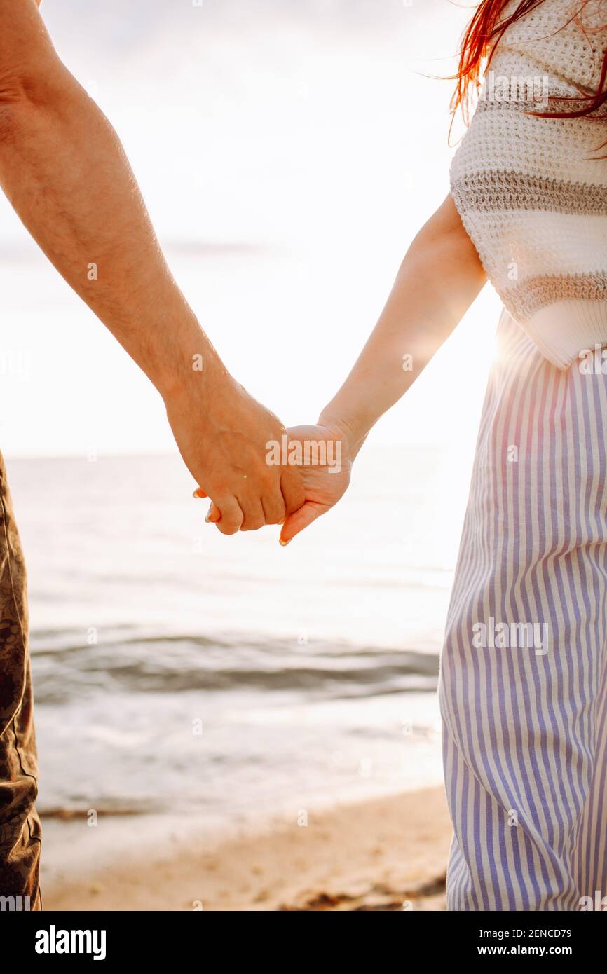 Unrecognizable couple hold their hands along empty ocean beach sand at sunset . Back side view. Stock Photo