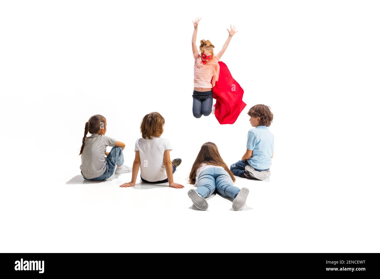 Flying. Child pretending to be a superhero with his friends sitting around  him. Kids excited and inspired by their brave friend in red coat isolated  on white background. Dreams, emotions concept Stock