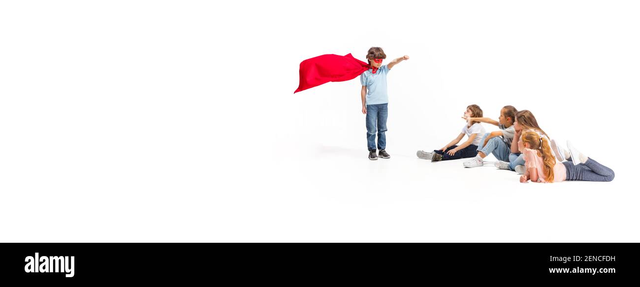 Flyer. Child pretending to be a superhero with his friends sitting around him. Kids excited and inspired by their brave friend in red coat isolated on white background. Dreams, emotions concept. Stock Photo