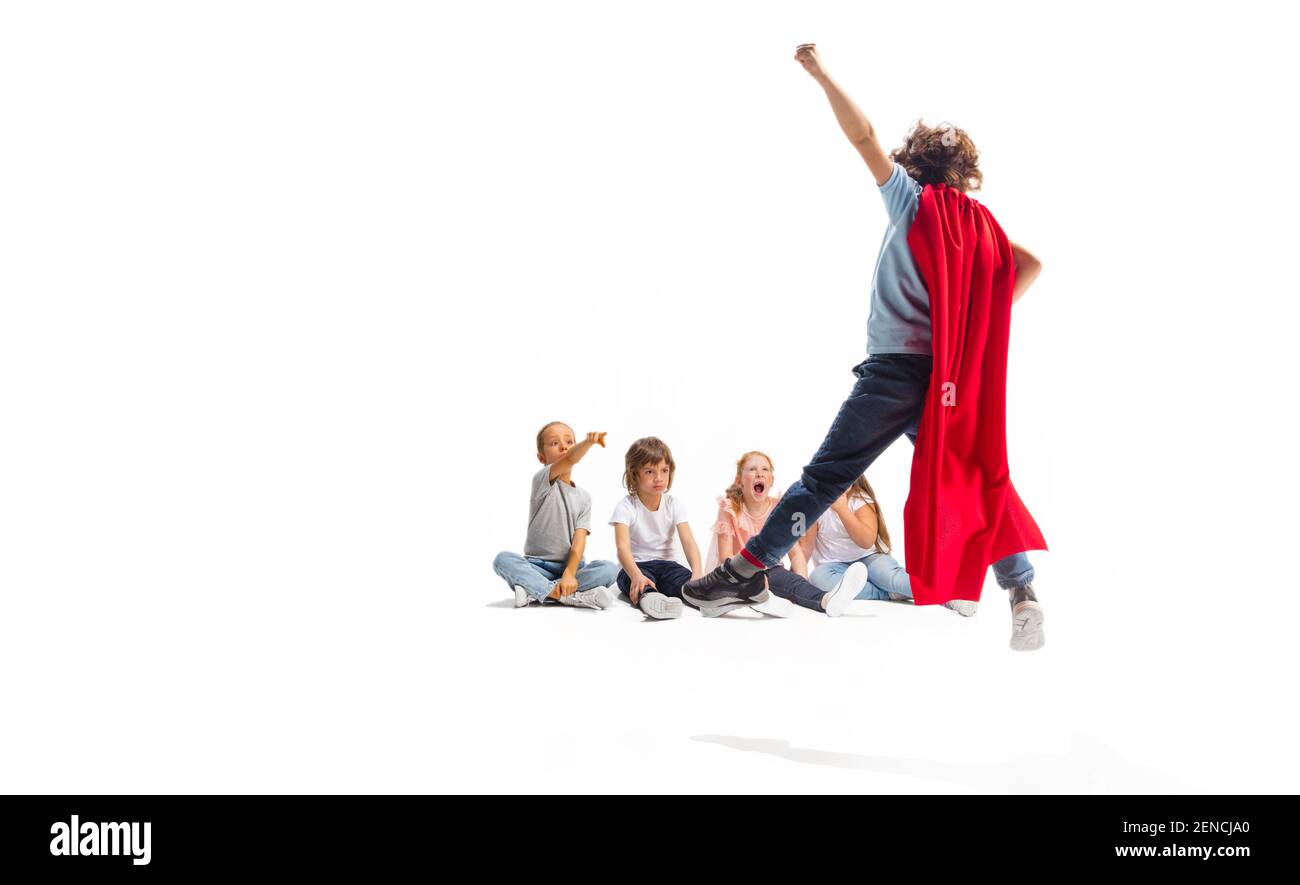 Power. Child pretending to be a superhero with his friends sitting around him. Kids excited, inspired by their strong friend in red coat isolated on white background. Dreams, emotions concept. Stock Photo