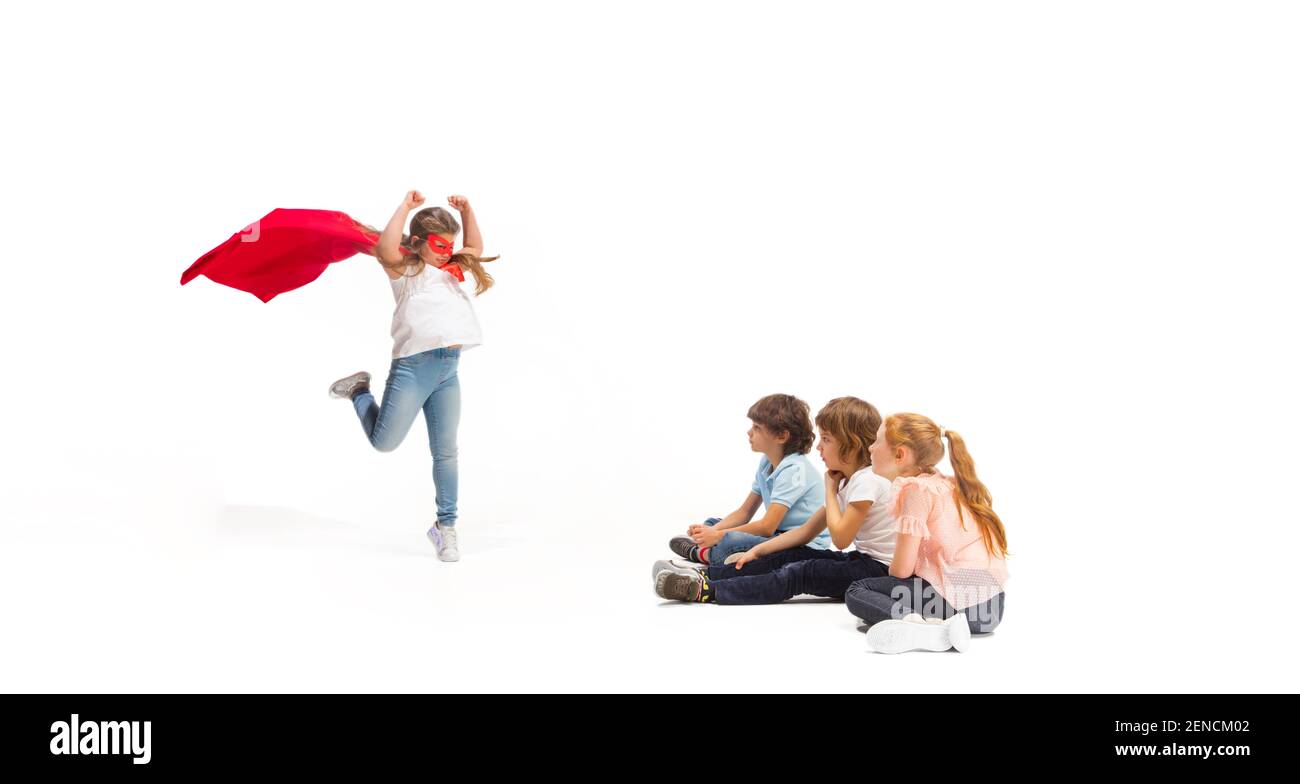 Flying. Child pretending to be a superhero with his friends sitting around  him. Kids excited and inspired by their brave friend in red coat isolated  on white background. Dreams, emotions concept Stock