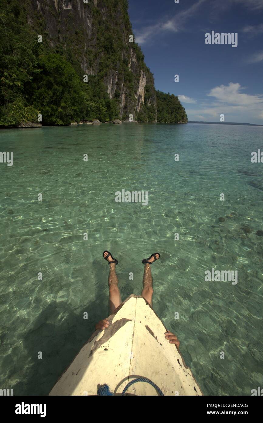 A man is having leisure time by floating on seawater while holding on to the foremost section of a boat on coastal water below cliff, one of the coastal tourism attractions close to Sawai ecotourism village in North Seram, Central Maluku, Maluku, Indonesia. Stock Photo