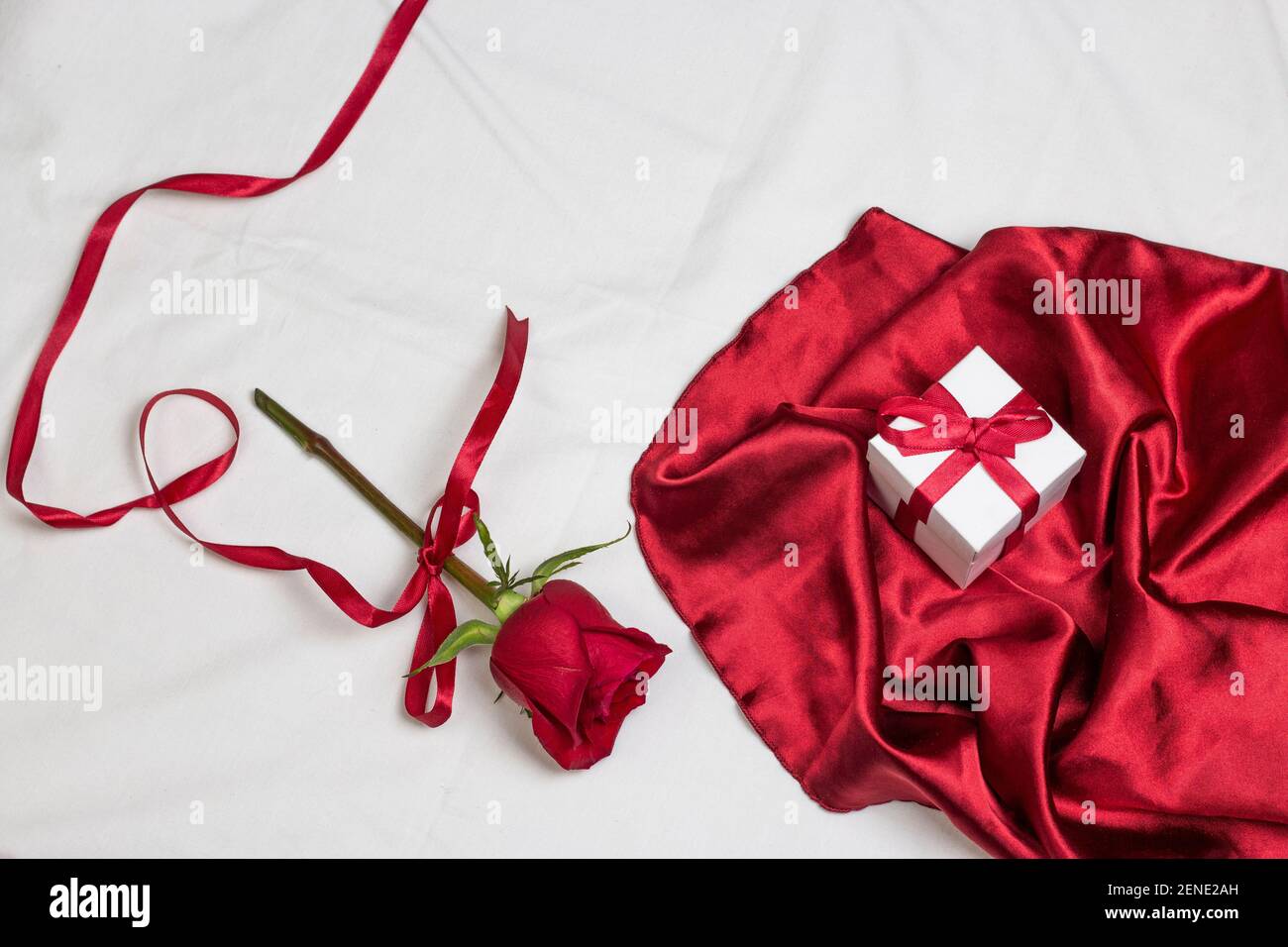 Red rose with red ribbon and red satin fabric with gif box on white bed with pillow. Valentine’s day or holiday romantic background Stock Photo