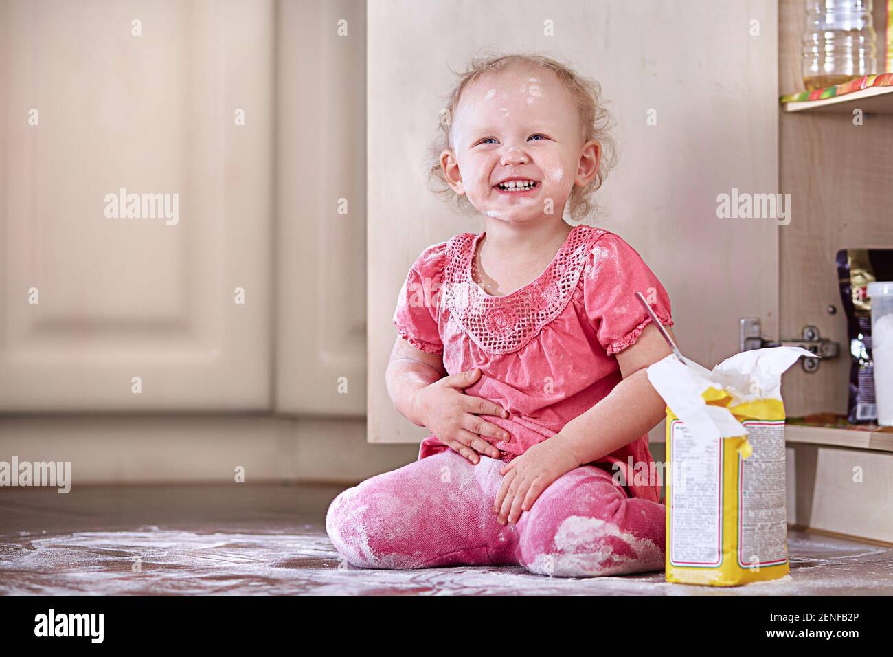 Playful laughing little baby girl smeared in flour sits on the kitchen floor. Copy space. Stock Photo