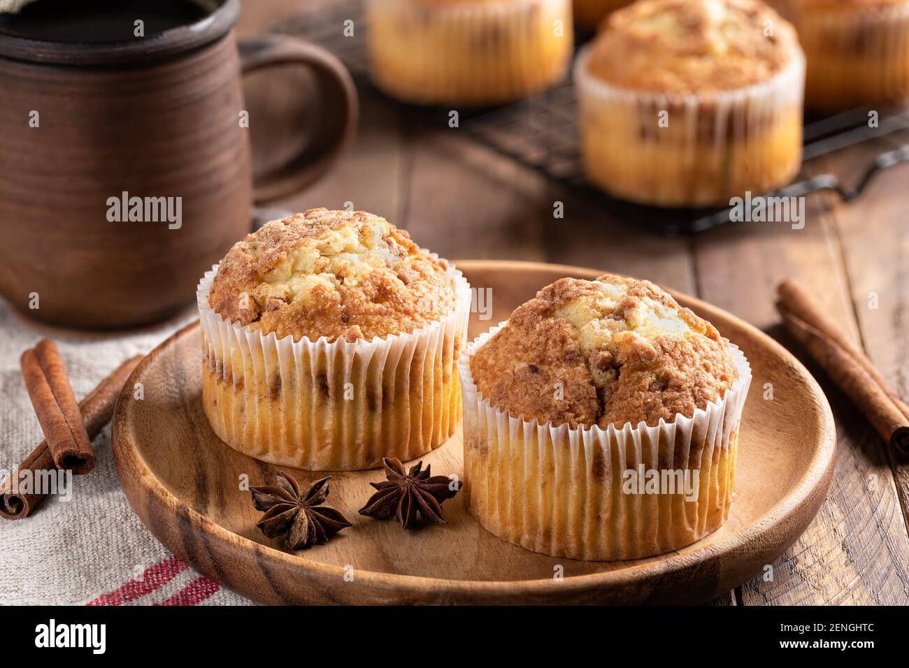 Two cinnamon muffins on a wooden plate with cup of coffee and muffins on a rack in background Stock Photo