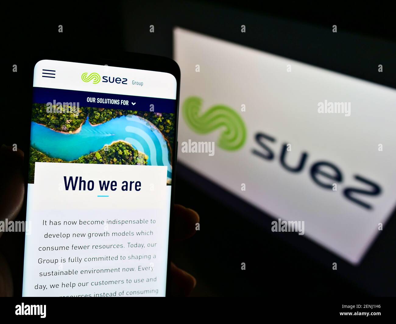 Person holding smartphone with website of French utility company Suez S.A. on screen in front of business logo. Focus on center-left phone display. Stock Photo