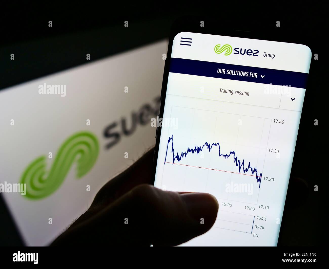 Person holding cellphone with website and chart of French utility company Suez S.A. on screen in front of logo. Focus on top-center of phone display. Stock Photo