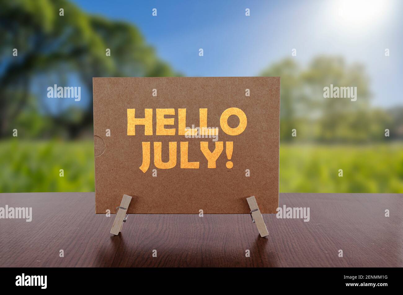 Hello july text on card on the table with sunny green park background. Stock Photo
