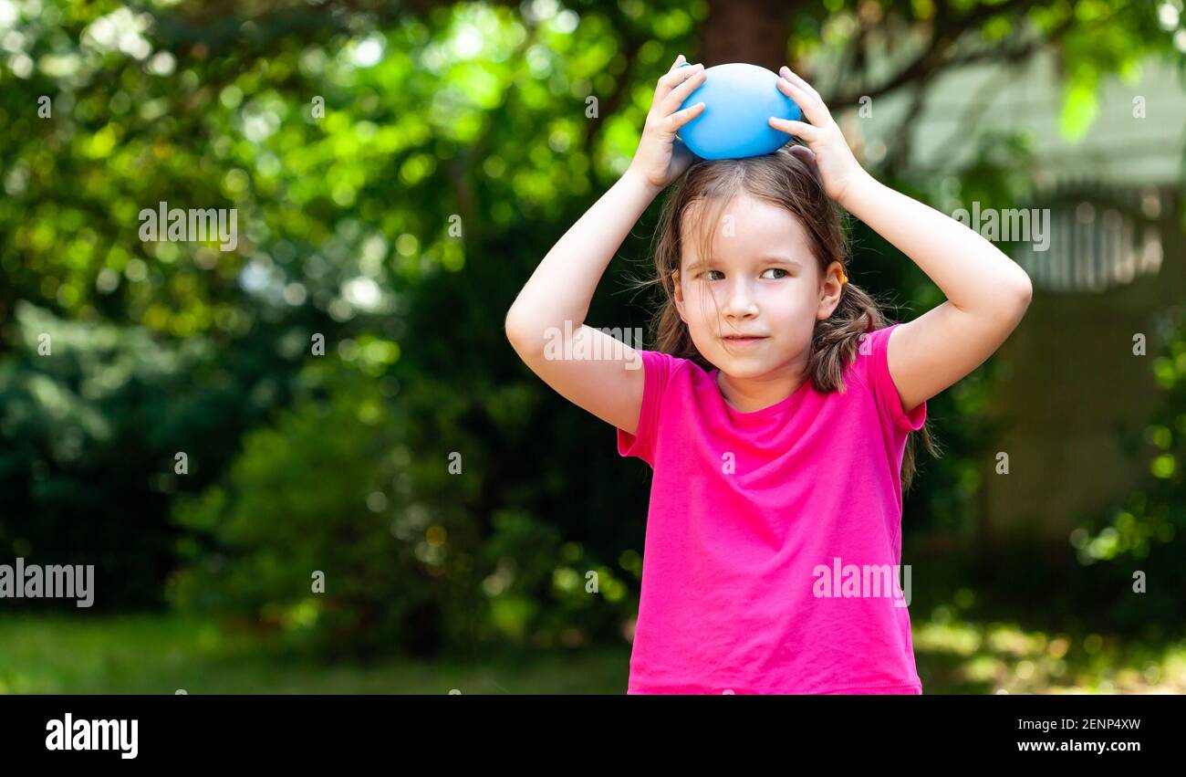 Little child, girl smiling playing with a water balloon outdoors, outside portrait, copy space. Kid holding a balloon filled with water over her head, Stock Photo