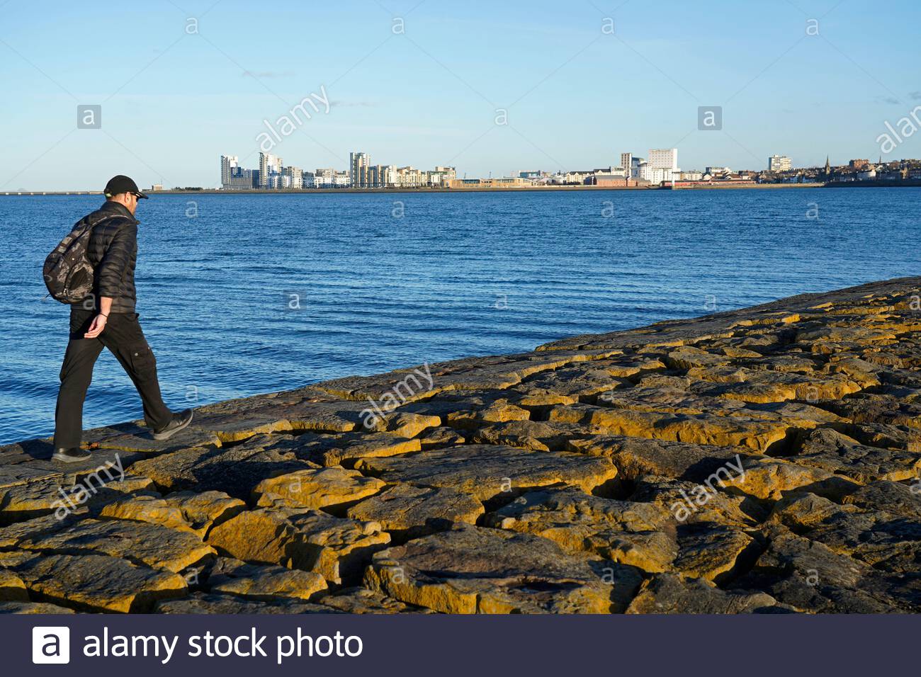 Edinburgh, Scotland, UK. 26th Feb 2021. Walking along the breakwater at Granton harbour with a view over the Forth Estuary on a cold and sunny late afternoon. View towards the modern development at Leith. Credit: Craig Brown/Alamy Live News Stock Photo