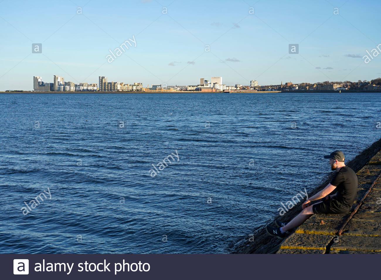 Edinburgh, Scotland, UK. 26th Feb 2021. Sitting on the breakwater at Granton harbour with a view over the Forth Estuary on a cold and sunny late afternoon. View towards the modern development at Leith.  Credit: Craig Brown/Alamy Live News Stock Photo