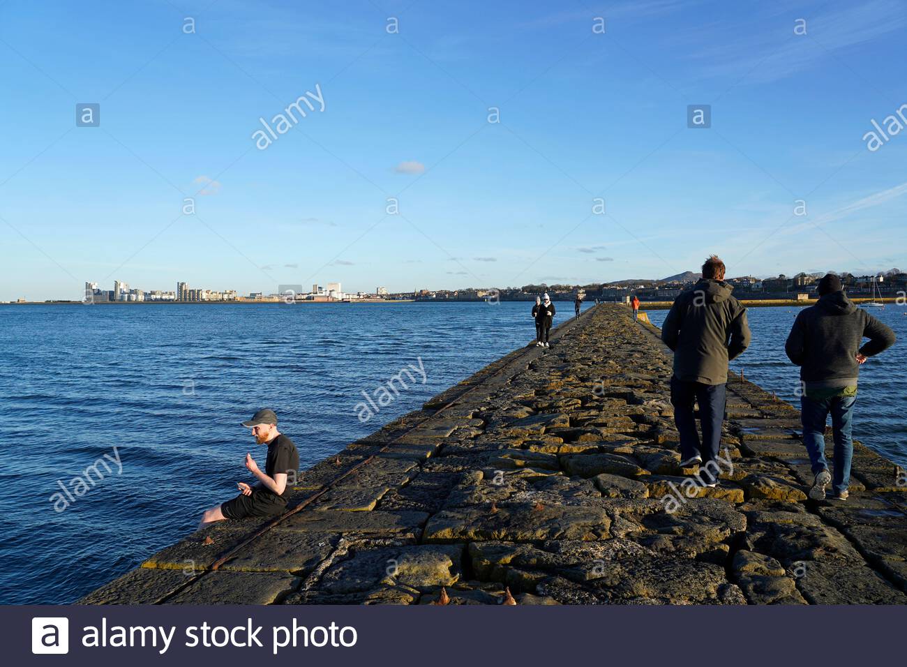 Edinburgh, Scotland, UK. 26th Feb 2021. People on the breakwater at Granton harbour with a view over the Forth Estuary on a cold and sunny late afternoon. View towards the modern development at Leith.  Credit: Craig Brown/Alamy Live News Stock Photo
