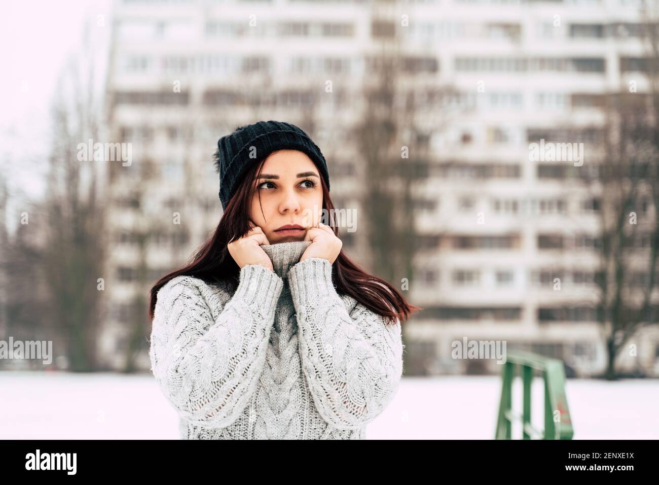 A pretty girl, dressed in a gray knitted sweater and a hat, covers her face with a sweater collar. Stock Photo