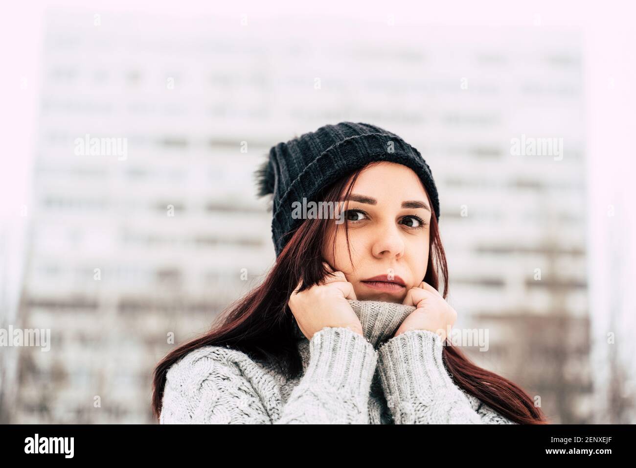 A pretty girl, dressed in a gray knitted sweater and a hat, covers her face with a sweater collar. Stock Photo