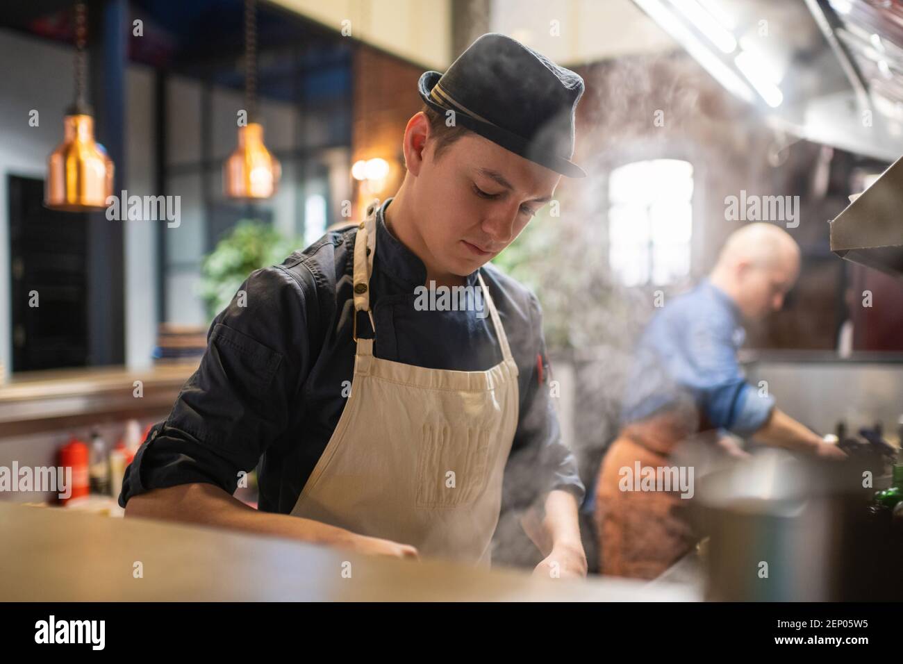 Young man in apron and hat preparing steamy dish in kitchen of cafeteria Stock Photo