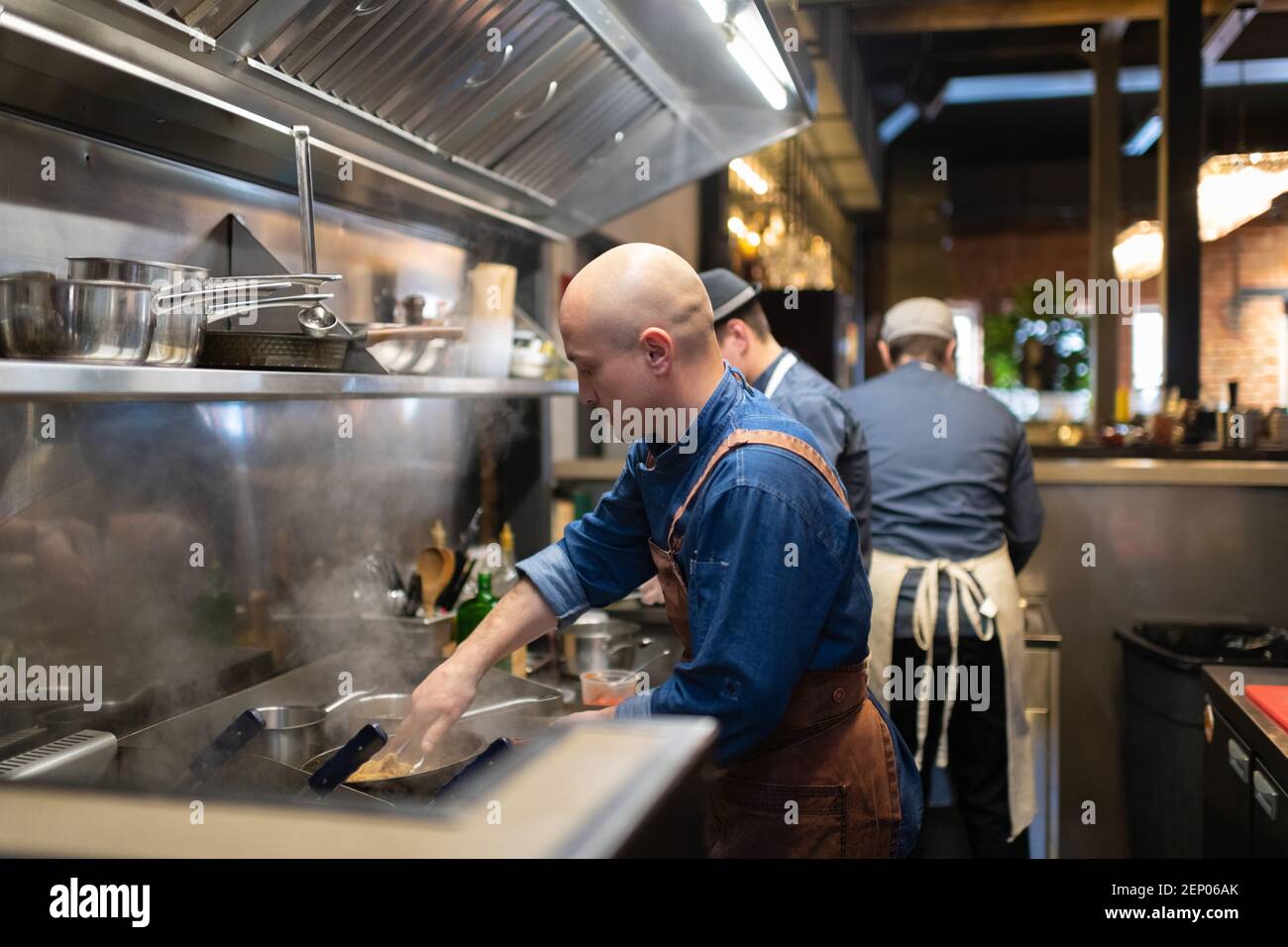 Bald man using tongs to mix pasta on frying pan while cooking in kitchen of cafe Stock Photo