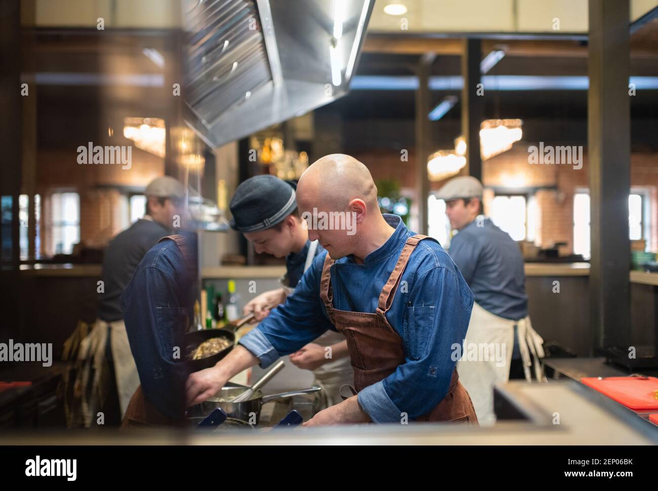 Bald man in apron boiling food near male employees during work in kitchen of cafe Stock Photo
