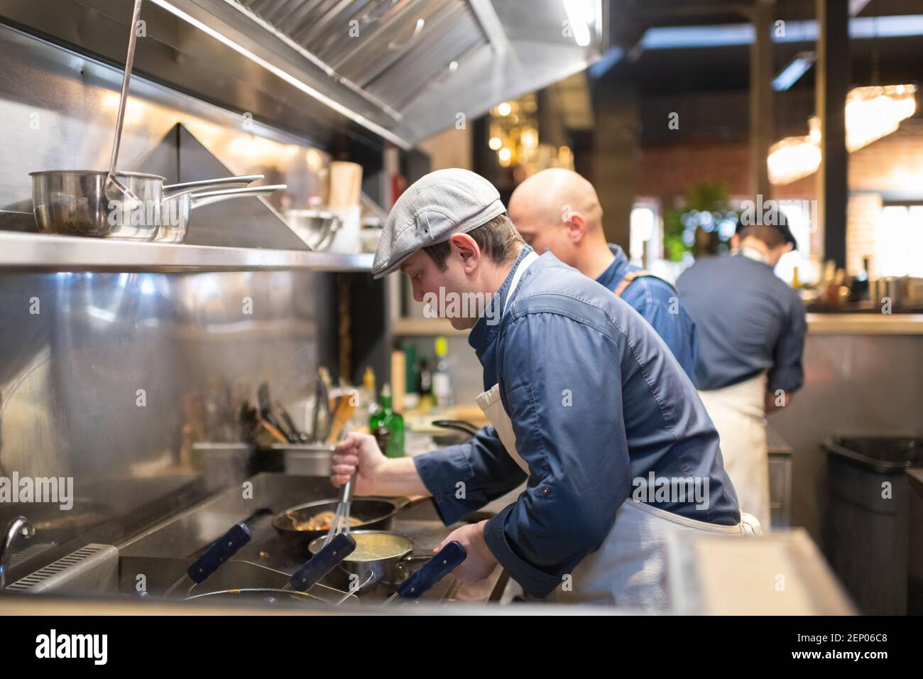 Man in cap whipping sauce in saucepan on stove while working in cafe kitchen Stock Photo