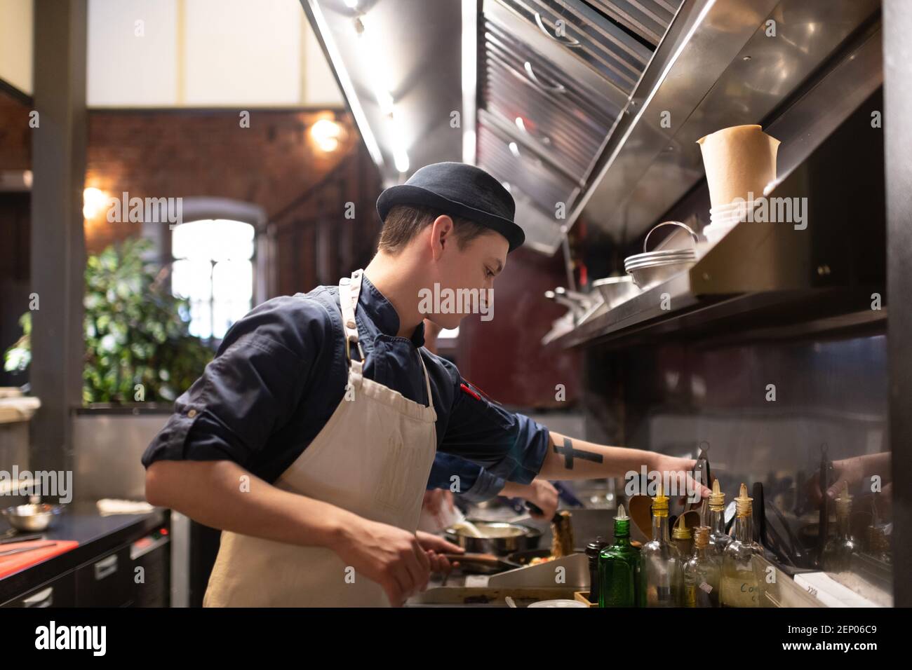 Young cook in hat and apron taking tongs while preparing dish in cafe kitchen Stock Photo