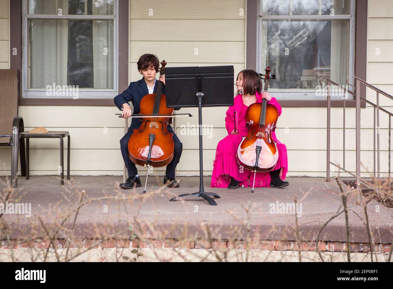 A tween boy sits on porch playing cello in suit while sister watches Stock Photo