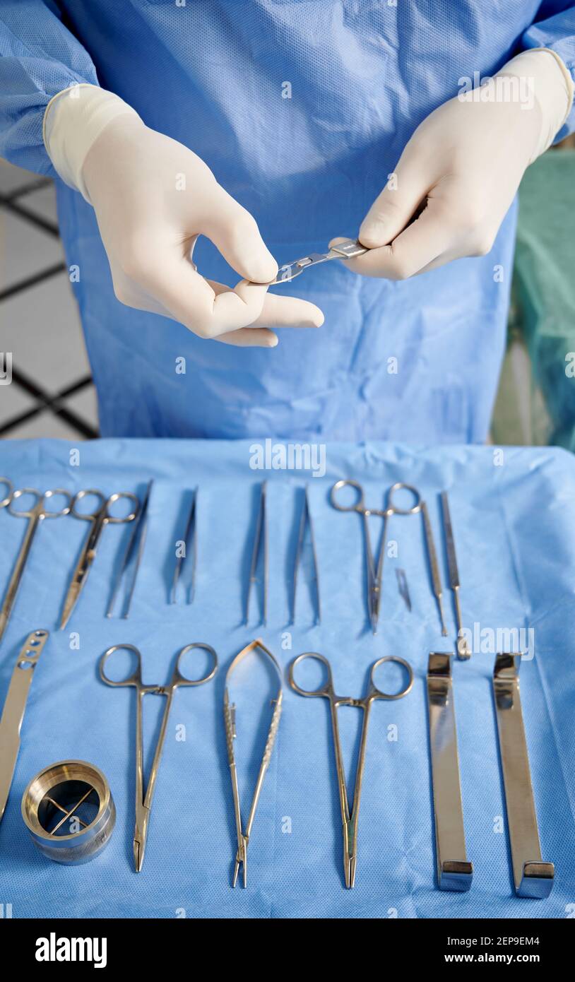 Doctor hands getting ready stainless steel medical scalpel for plastic surgery with set of various tools and instruments on table. Surgeon in white sterile gloves. Concept of medicine. Stock Photo