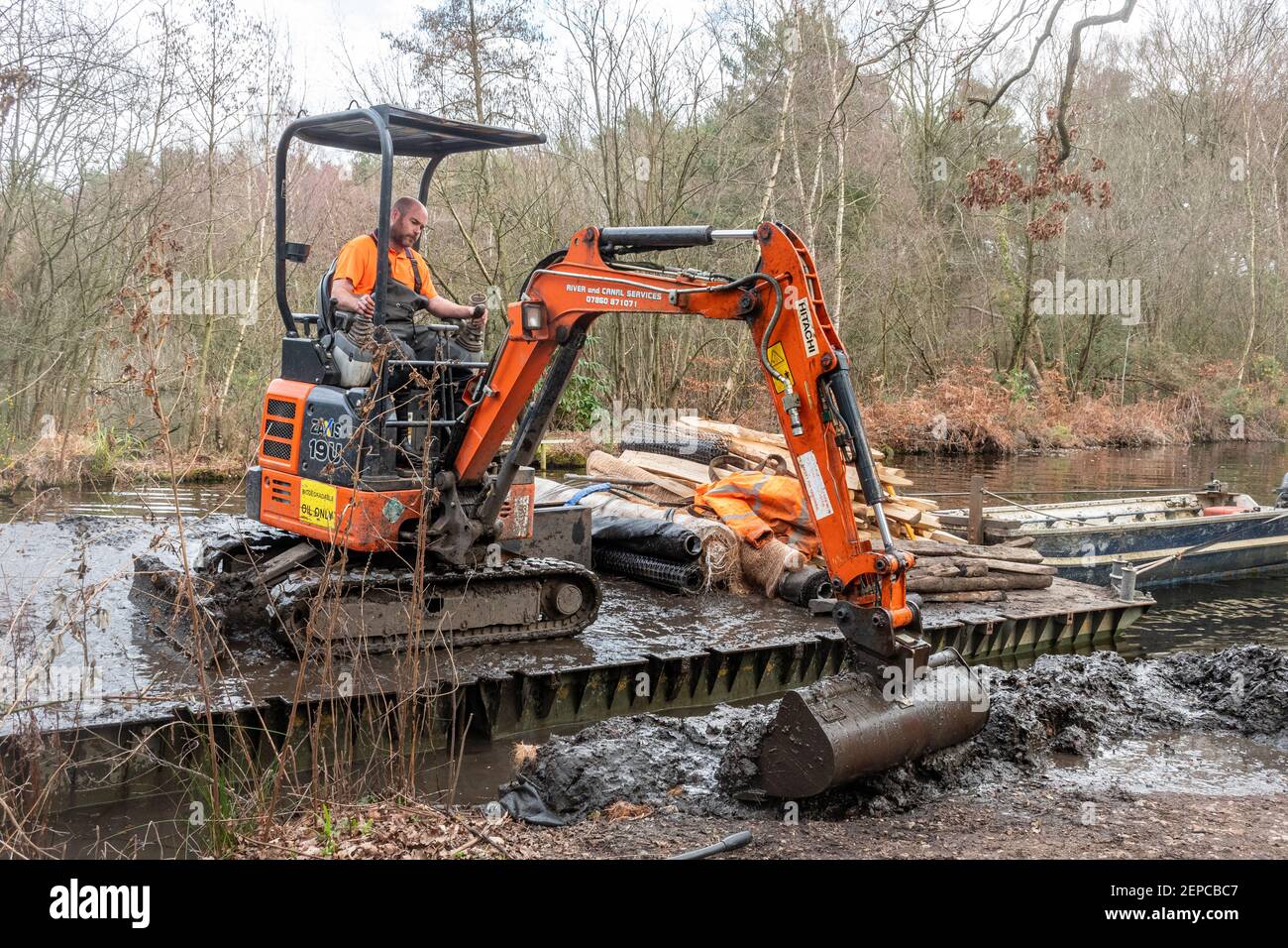 Man working in an excavator or digger on a floating work pontoon repairing an eroded canal bank on Basingstoke Canal in Surrey, UK Stock Photo