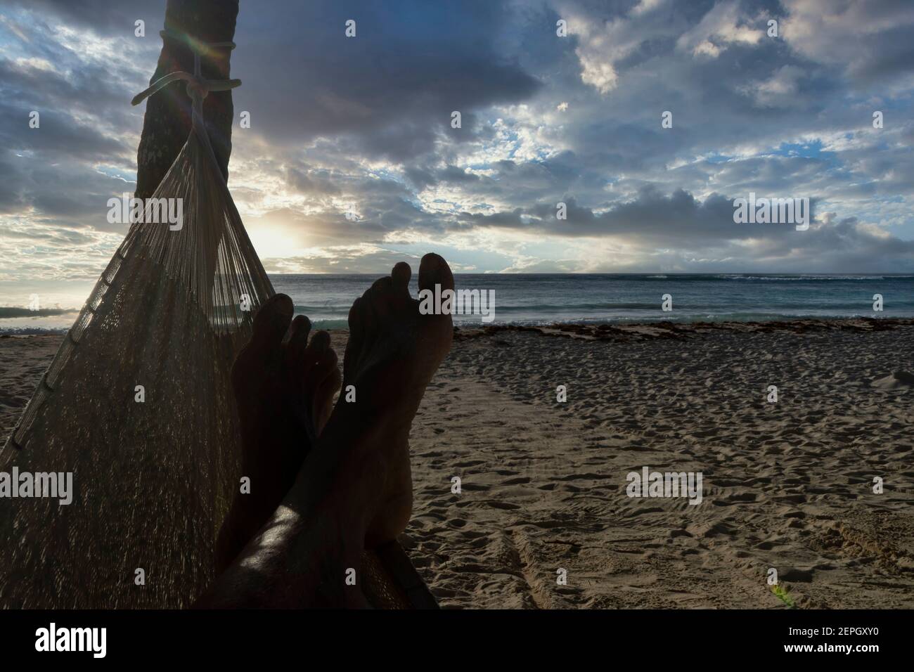 Close-up of a tourist's feet relaxing while looking at the sea, in a hammock on a tropical Mexican beach at sunrise. In the background the cloudy sky. Stock Photo