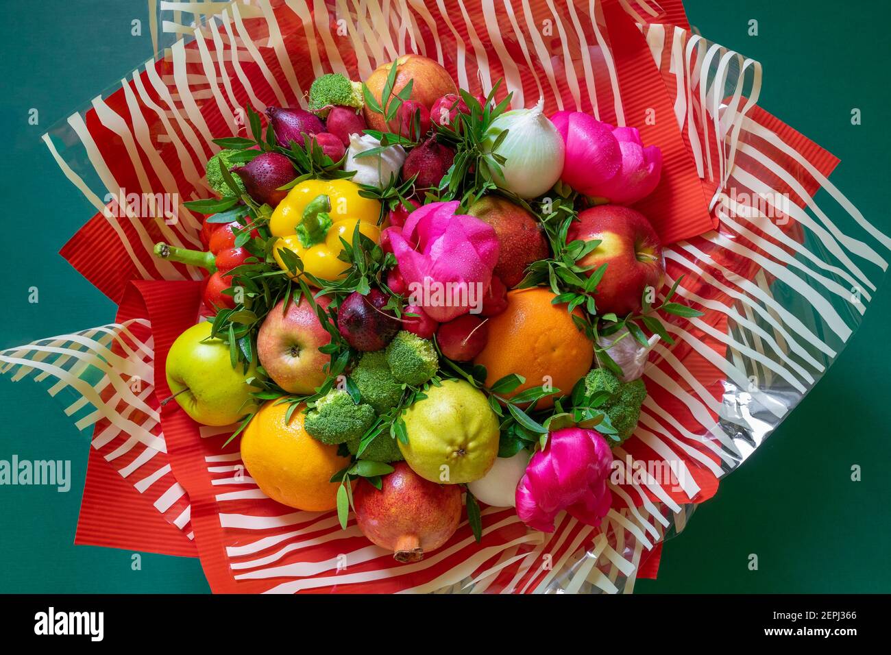 A vibrant multicolored bouquet of vegetables and fruits with pink peonies and green leaves wrapped in corrugated red paper and striped clear cellophan Stock Photo