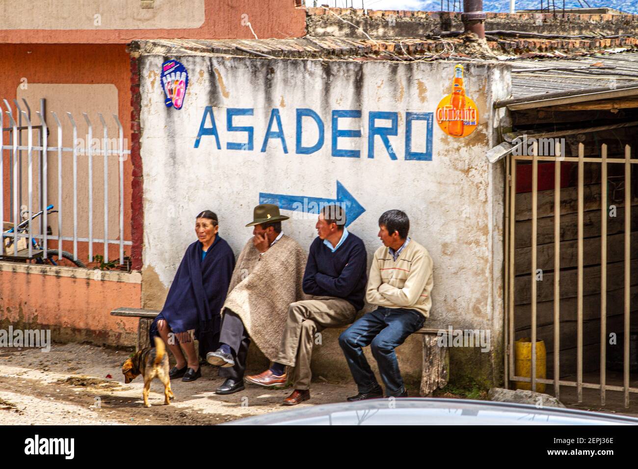 1 dog,Colombian 3 men, 1 women sitting on bench, waiting. 1 in traditional hat  poncho, Ruana. Asadero sign Boyacá Department, Colombia, South America Stock Photo