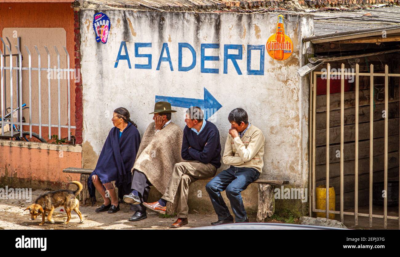 1 dog,Colombian 3 men, 1 women sitting on bench, waiting. 1 in traditional hat  poncho, Ruana. Asadero sign Boyacá Department, Colombia, South America Stock Photo