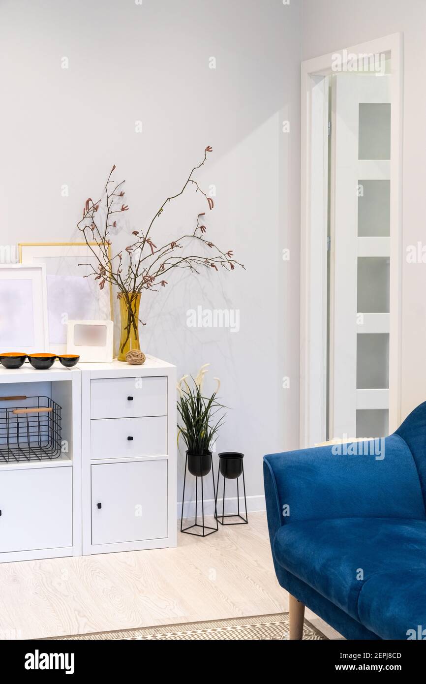 A fragment of the interior of a small cozy light-lined living space. Blue comfortable sofa, white cabinet on which are placed vases with twigs Stock Photo