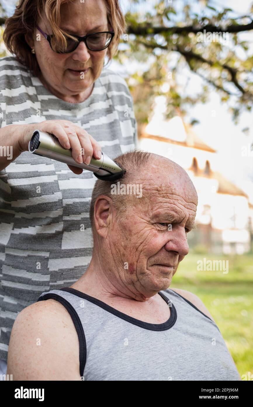 Woman cutting hair with electric razor to senior man outdoors at garden. Elderly people take care each other. Happy senior couple Stock Photo