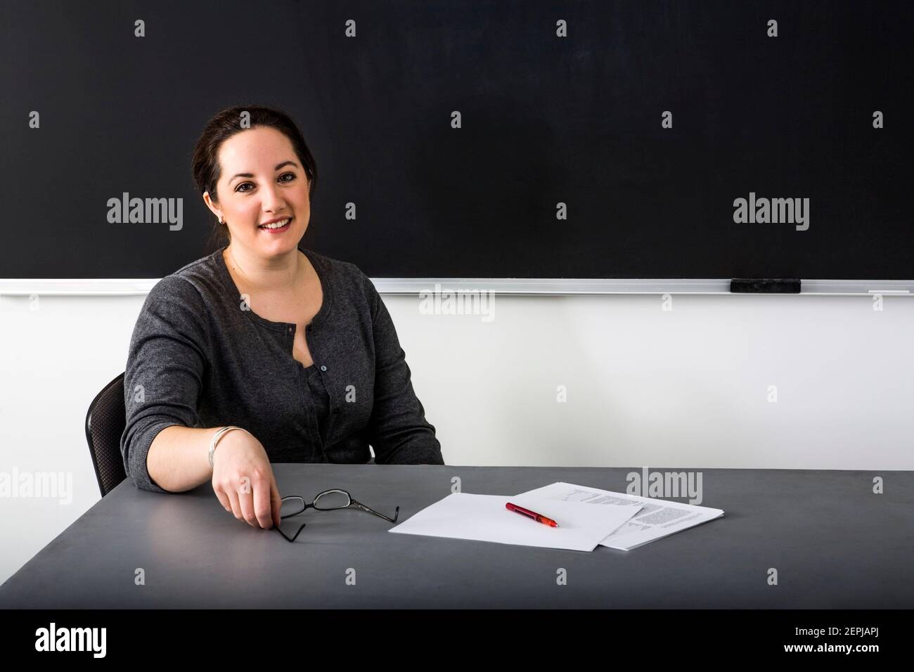 A young teacher sits at her desk in front of a blackboard. Stock Photo