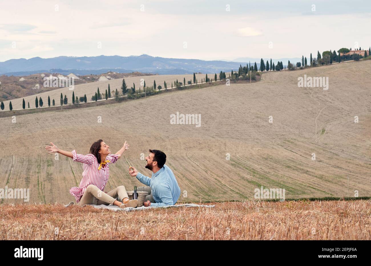 A young cheerful couple is delighted by a large meadow where they have a picnic on a beautiful day. Love, relationship, picnic, together Stock Photo