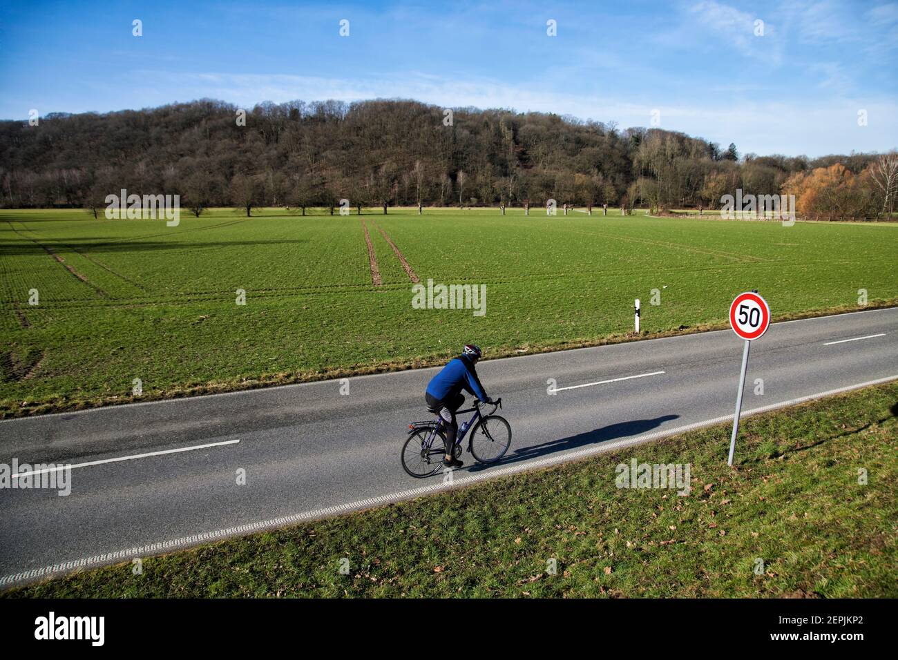 Fahrradfahrer auf einer zweispurigen Landstrasse vor einem Feld. Im Hintergrund befindet sich ein bewaldeter Huegel. Es ist Winter. Zu sehen ist ein V Stock Photo