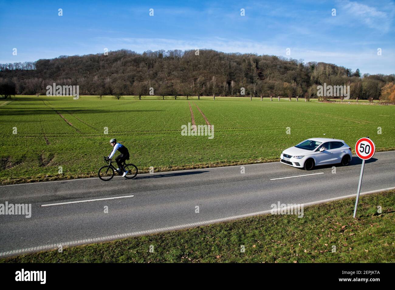 Fahrradfahrer auf einer zweispurigen Landstrasse vor einem Feld. Im Hintergrund befindet sich ein bewaldeter Huegel. Es ist Winter. Hinter dem Fahrrad Stock Photo