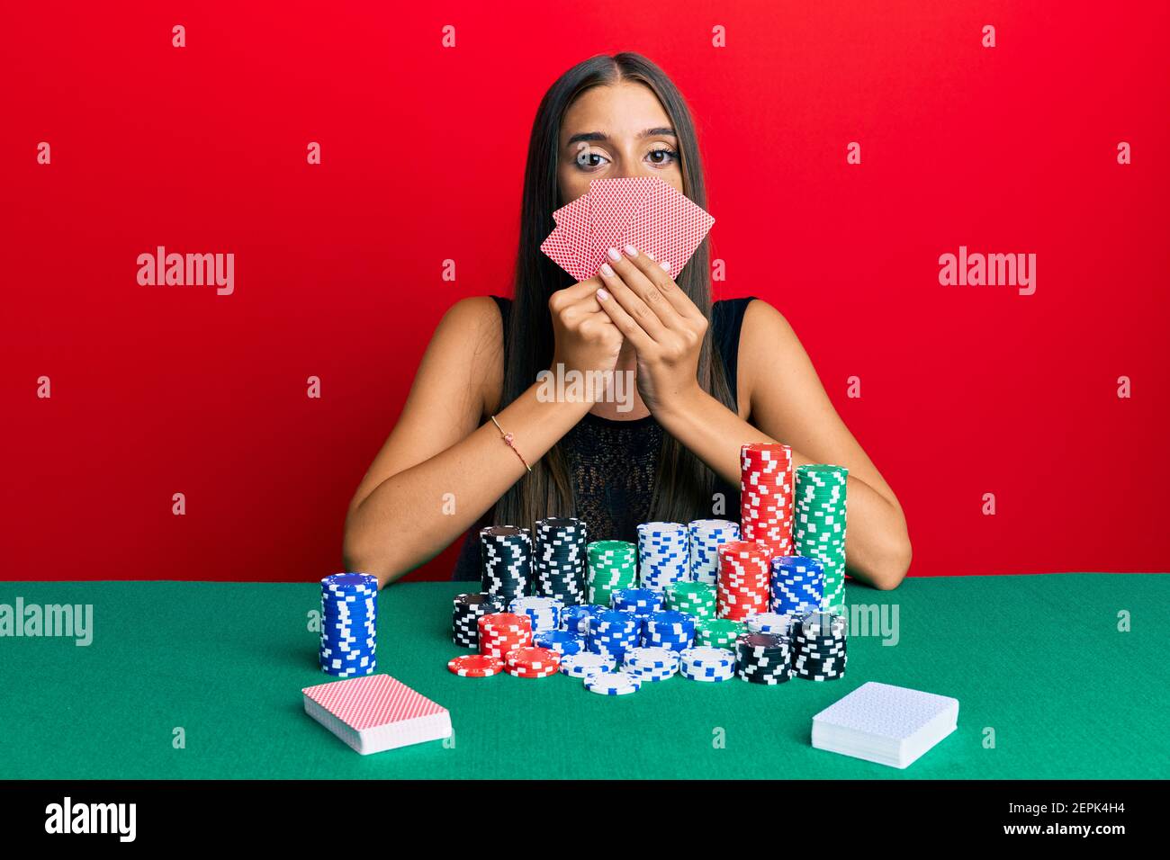 Young hispanic woman playing gambling poker covering face with cards relaxed with serious expression on face. simple and natural looking at the camera Stock Photo