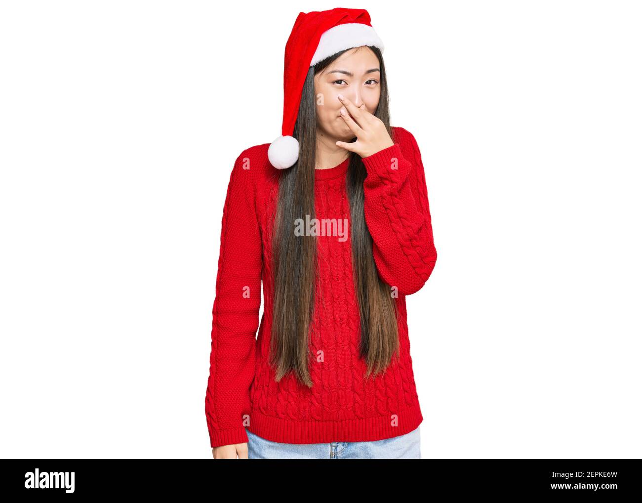 Young chinese woman wearing christmas hat smelling something stinky and disgusting, intolerable smell, holding breath with fingers on nose. bad smell Stock Photo