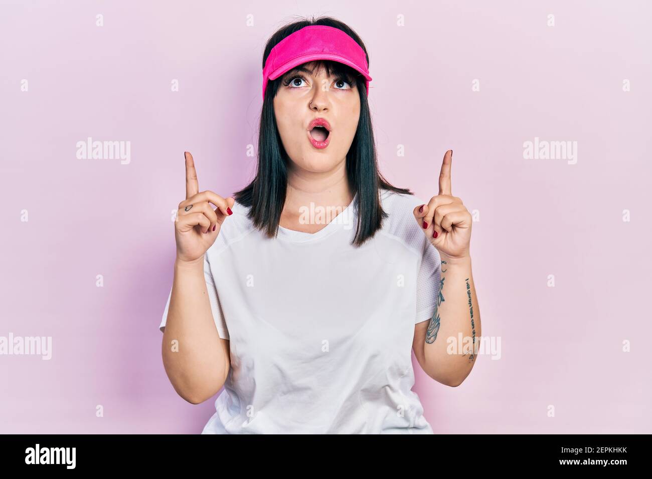 Young hispanic woman wearing sportswear and sun visor cap amazed and surprised looking up and pointing with fingers and raised arms. Stock Photo