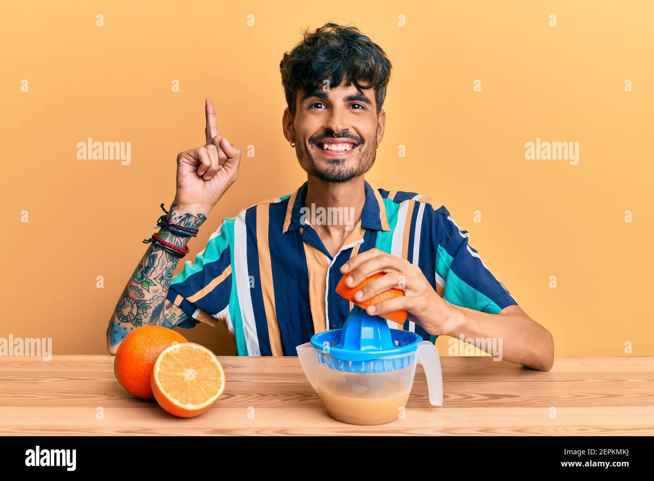 Young hispanic man sitting on the table using juicer surprised with an