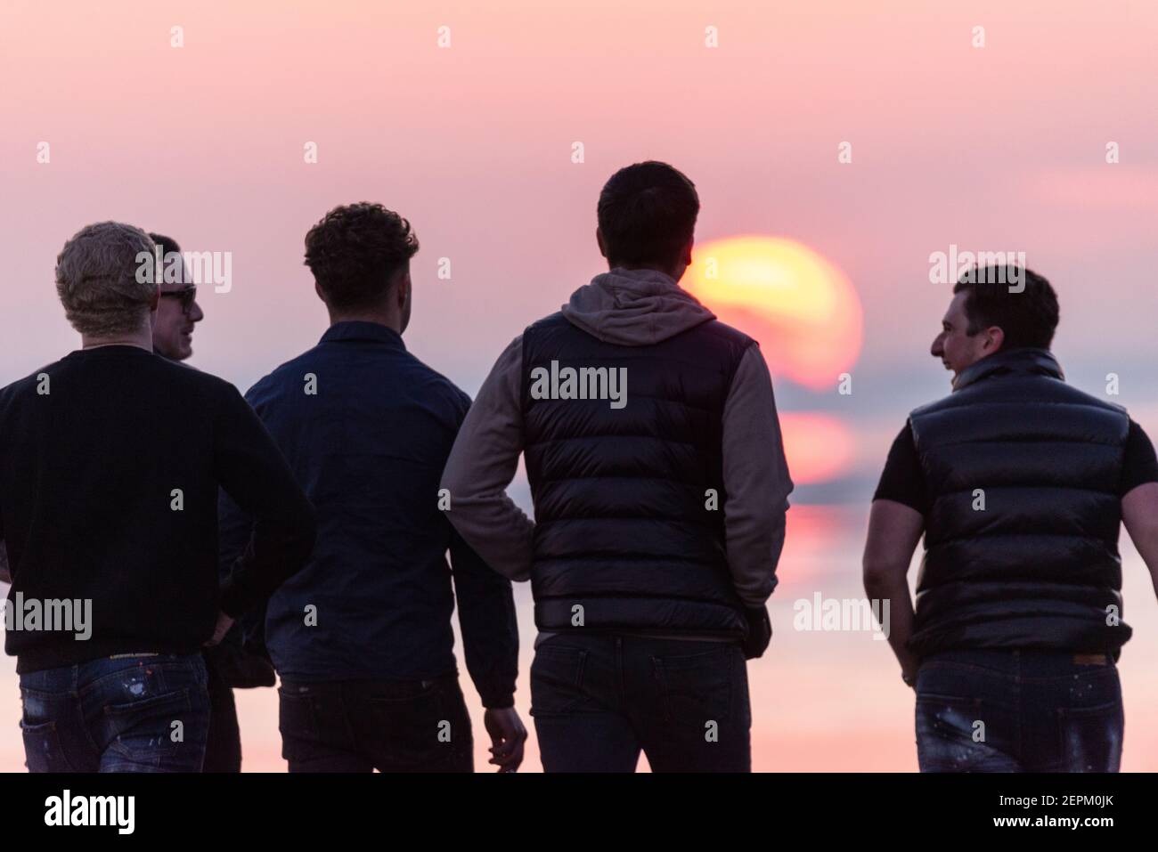Group of males walking on Southend on Sea seafront at sunset during COVID 19 lockdown. Sun setting behind people out walking in evening Stock Photo