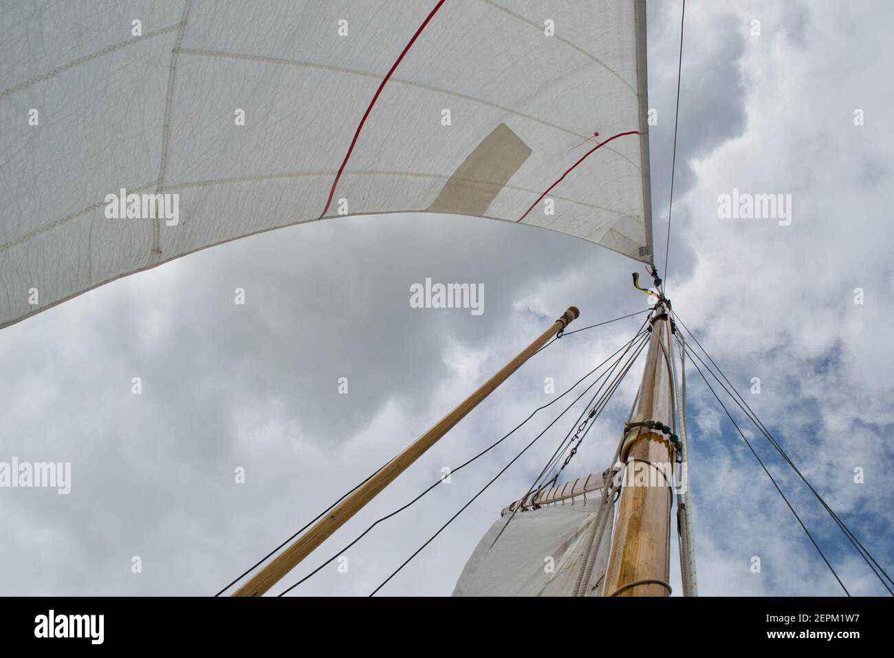 Looking up the mast of a gaff rigged sailing yacht: white sails, wooden mast and gaff, rope shrouds, halyards and sheets, and blue cloudy sunny sky Stock Photo