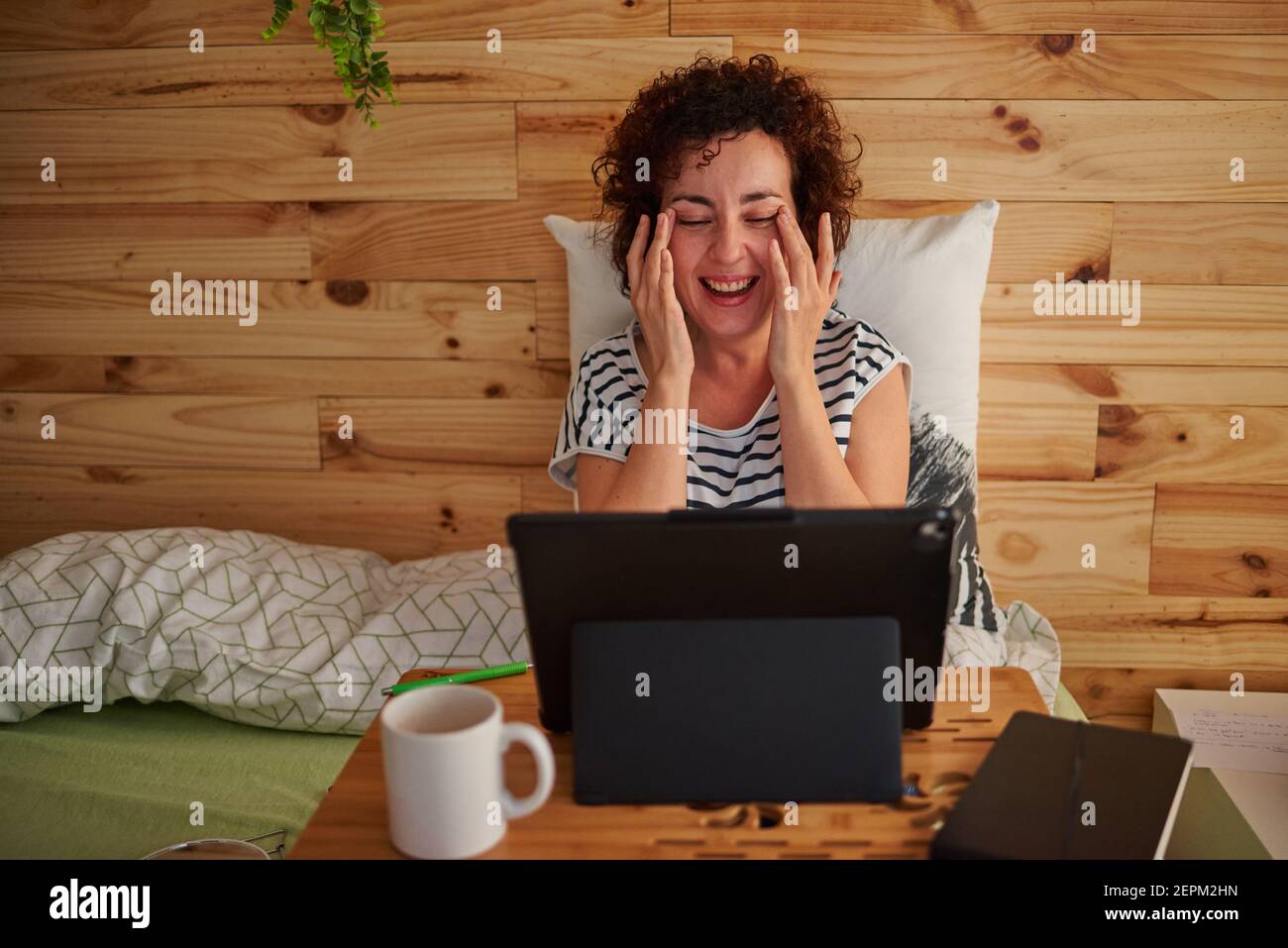 A curly-haired Caucasian woman wiped away tears from laughing so much. She is sitting on the bed with a folding table that serves as her desk. On the Stock Photo
