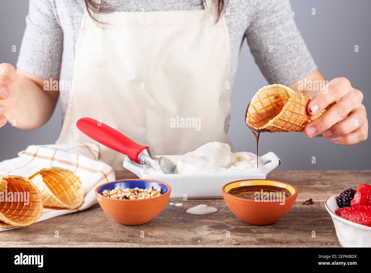 A woman chef is dipping homemade waffle ice cream cones into chocolate melt and walnut pieces for artisan look. Other hand rolled cones are there. A m Stock Photo