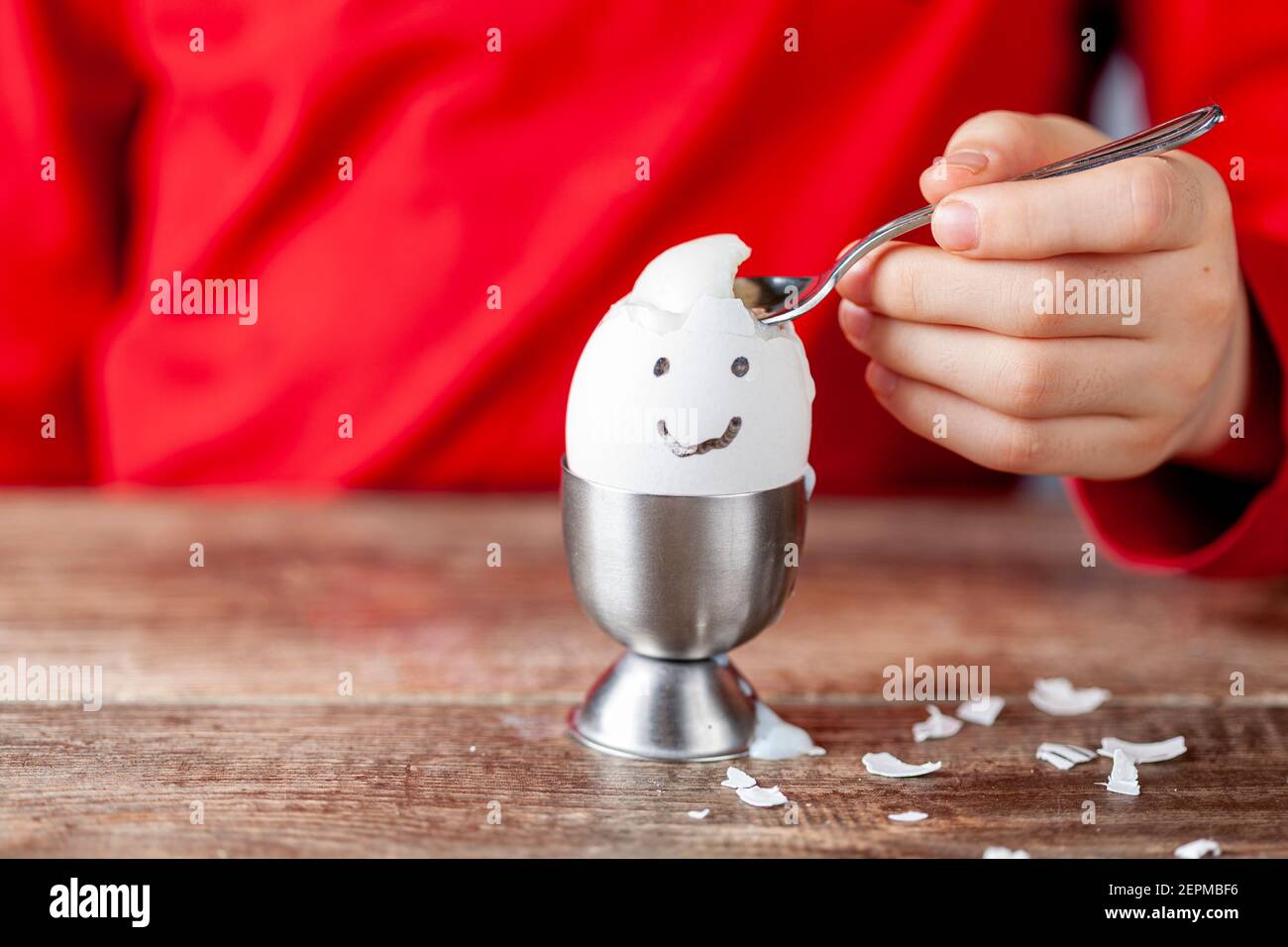 A small child is eating soft boiled humpty dumpty egg placed in egg cup on a wooden breakfast table. She uses spoon to eat the egg from its shell. She Stock Photo