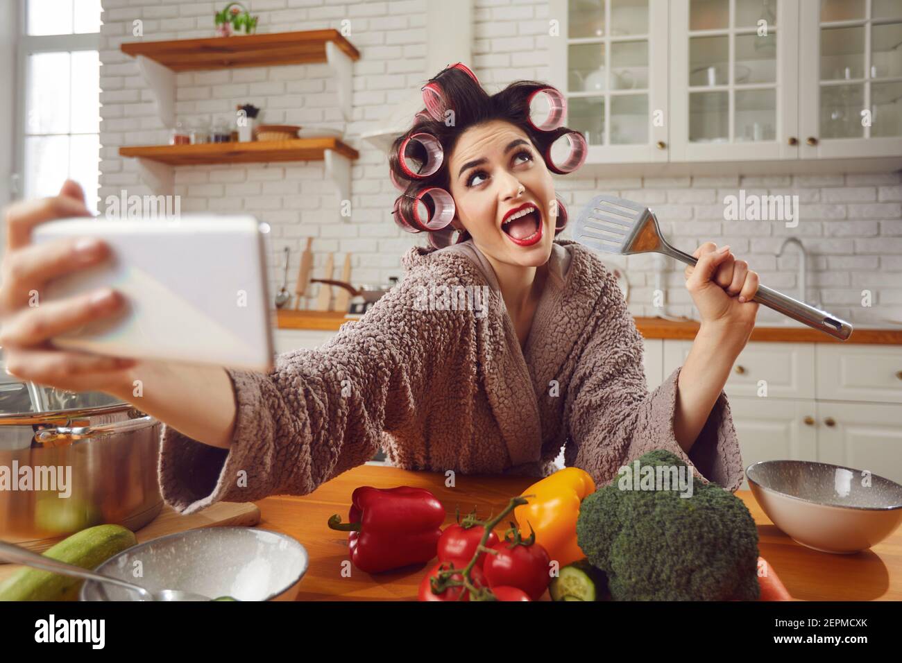 Woman in hair curlers taking funny selfie on mobile while cooking in the kitchen Stock Photo