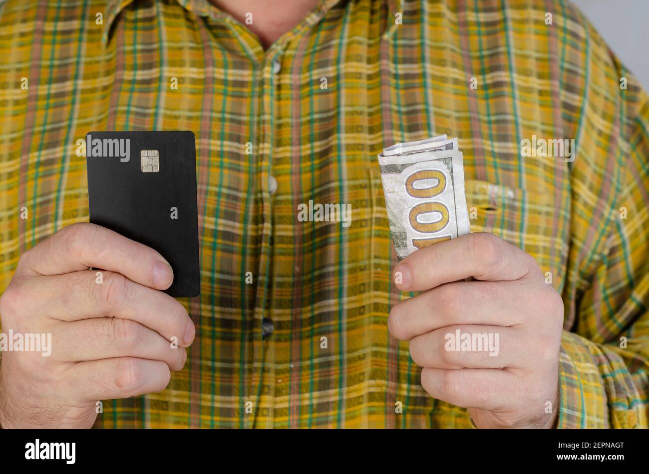 An adult man in a green and yellow plaid shirt holds money and a bank card at chest level. Male hands with a one hundred dollar bill and a black credi Stock Photo