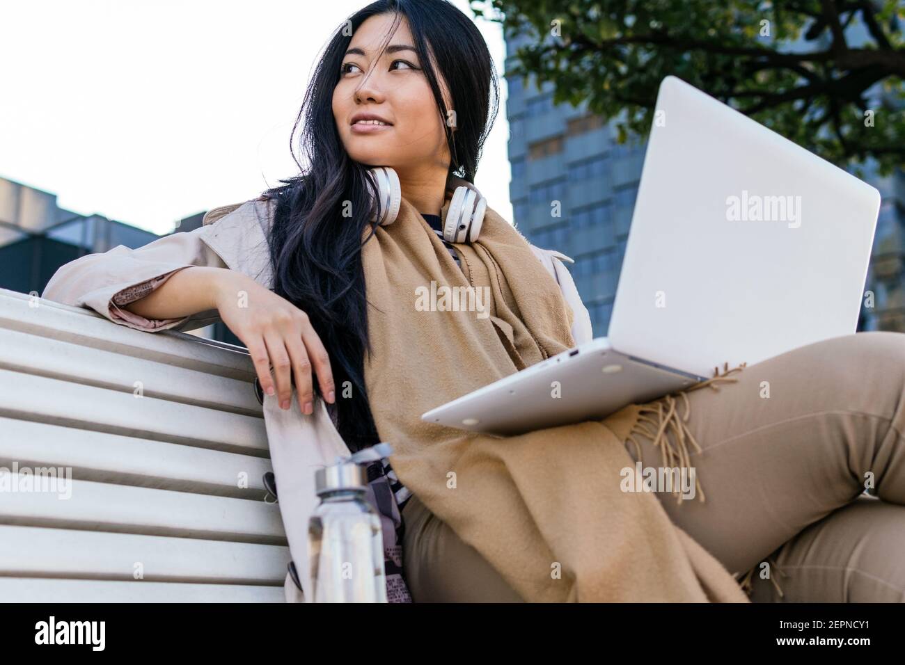 Ethnic young Asian female wearing warm outerwear browsing netbook while working remotely sitting on a bench on the street Stock Photo